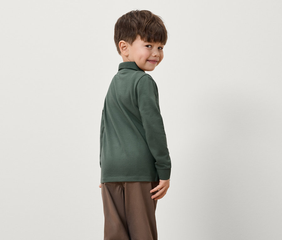 Ein Junge schaut über seine Schulter und trägt ein grünes Langarm-Poloshirt und braune Hosen.