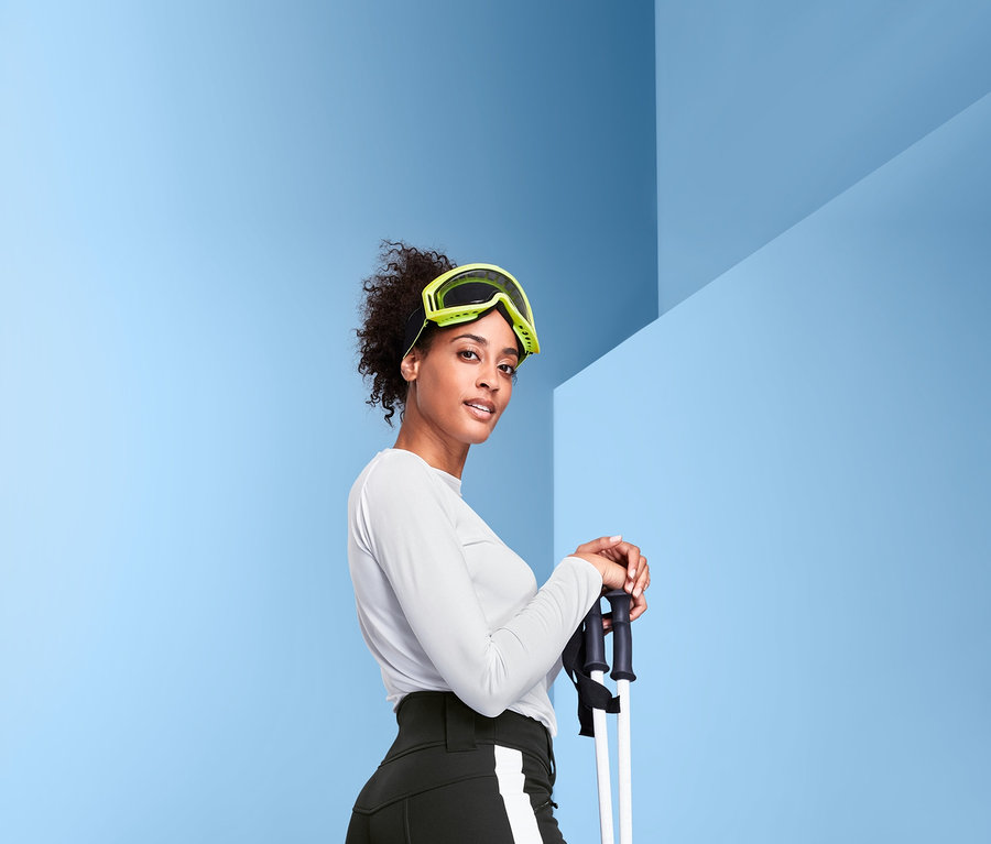 Frau mit Skibrille hält Skistöcke und trägt eine Jetskihose und Skischuhe.