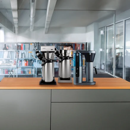 Drei Kaffeemaschinen stehen auf einer Holzplatte, im Hintergrund eine Bibliothek und Glaswände eines Büros.