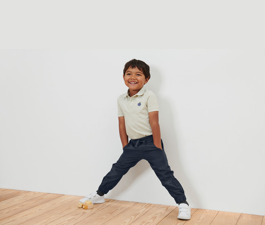 Ein lächelnder Junge in einem beigen Poloshirt, dunkelblauen Hosen und weißen Schuhen steht an einer weißen Wand.