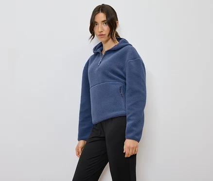 Frau posiert in einem blauen Fleecepullover mit Kapuze und schwarzen Hosen.