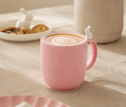 Auf einem Tisch steht eine rosa Tasse mit einem weißen Hasen am Rand, gefüllt mit einem Getränk mit Latte Art. Im Hintergrund befinden sich eine weiße Vase mit Blumen, ein Teller mit Keksen und eine Hasenfigur.