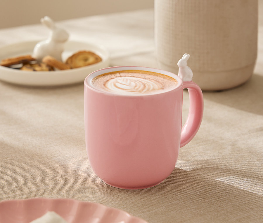Auf einem Tisch steht eine rosa Tasse mit einem weißen Hasen am Rand, gefüllt mit einem Getränk mit Latte Art. Im Hintergrund befinden sich eine weiße Vase mit Blumen, ein Teller mit Keksen und eine Hasenfigur.