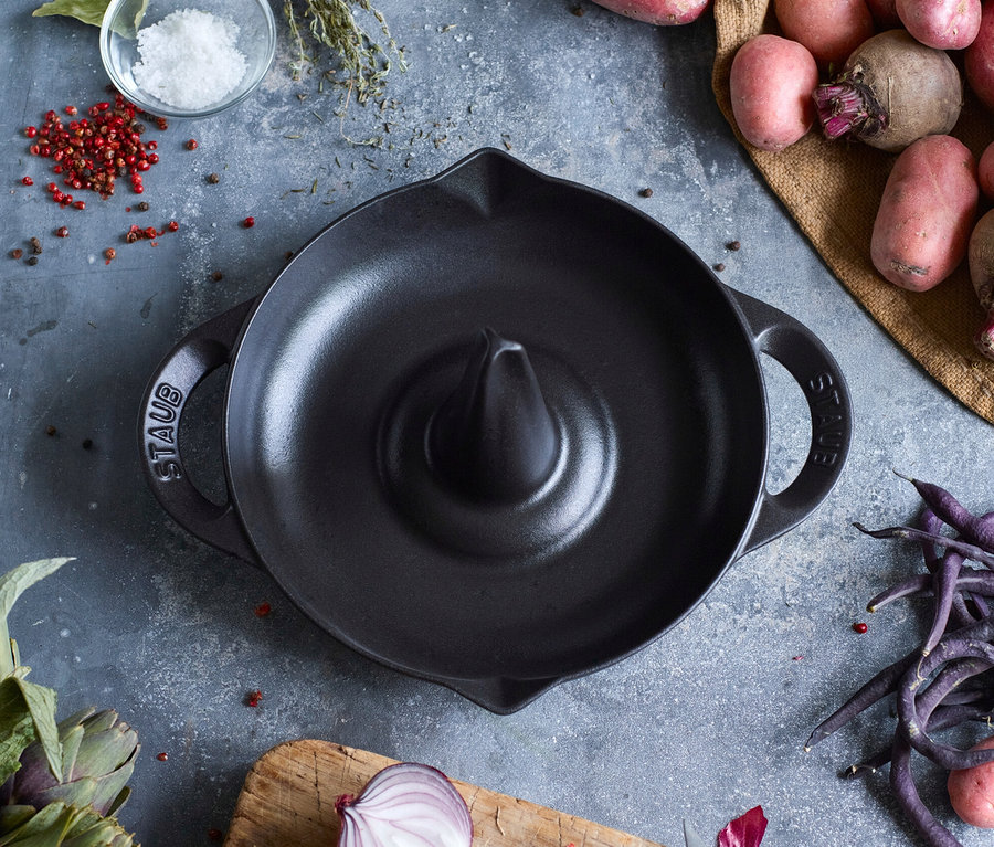 Schwarzer STAUB Hähnchen-Bräter umgeben von Zutaten wie Kartoffeln, Zwiebeln und Kräutern.