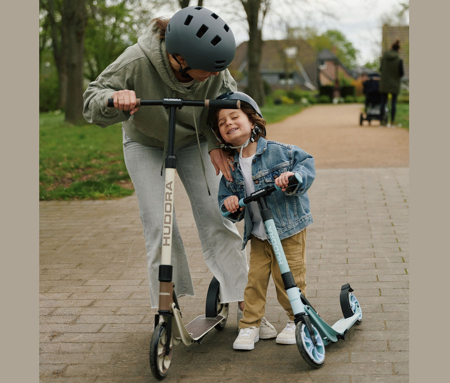 Eine Frau im grauen Hoodie und ein Junge auf HUDORA Scooter »BigWheel 180 Advanced Junior« Rollern auf einem Gehweg.