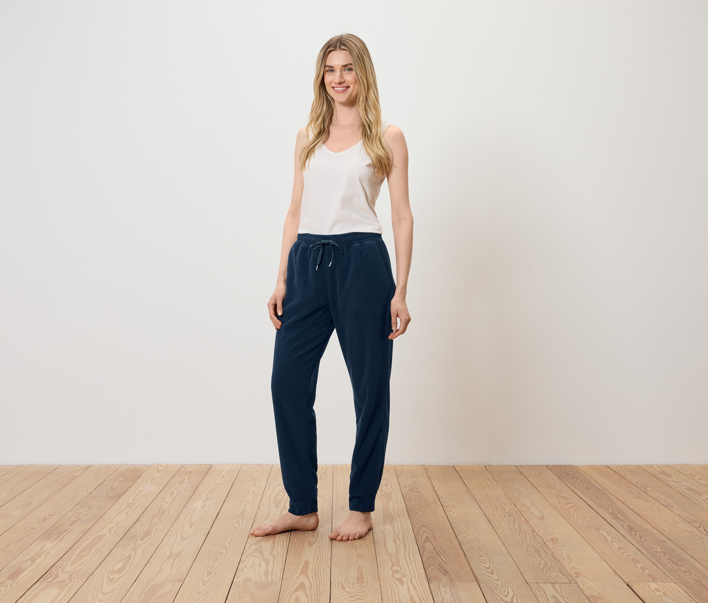 Eine Frau steht auf einem Holzboden und trägt ein weißes Top und eine dunkelblaue Trainingshose.