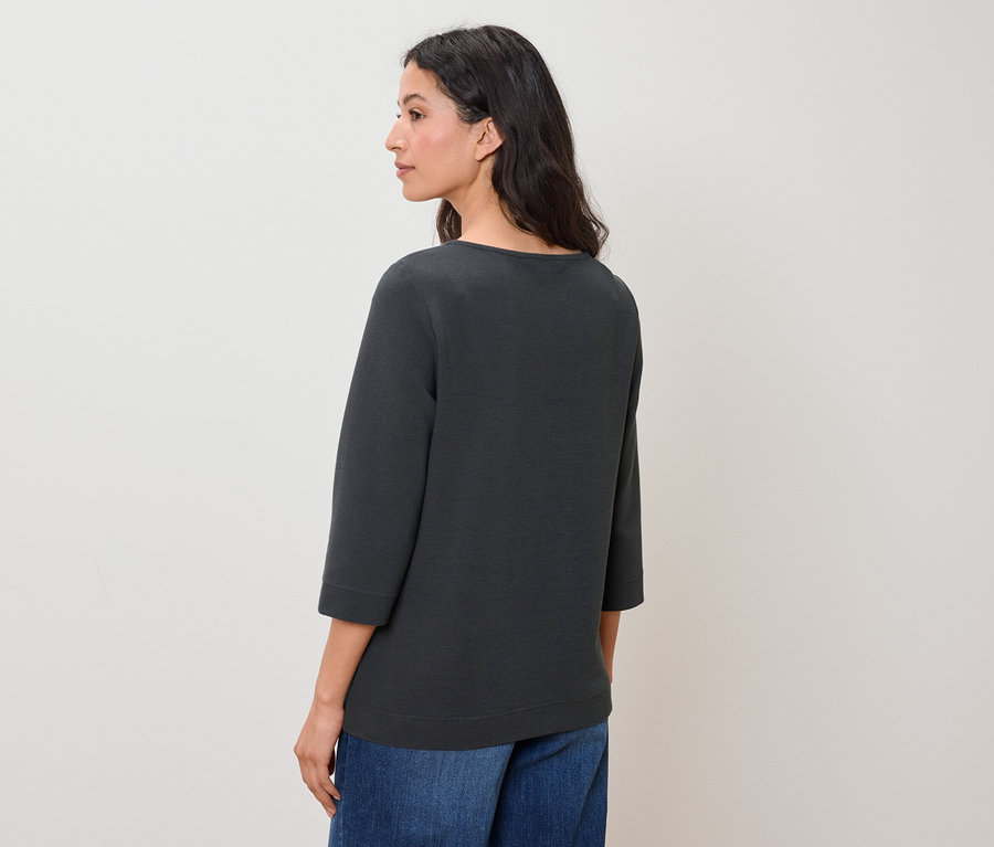 Eine Frau schaut nach links und trägt blaue Jeans und ein graugrünes Oberteil mit Dreiviertelärmeln.