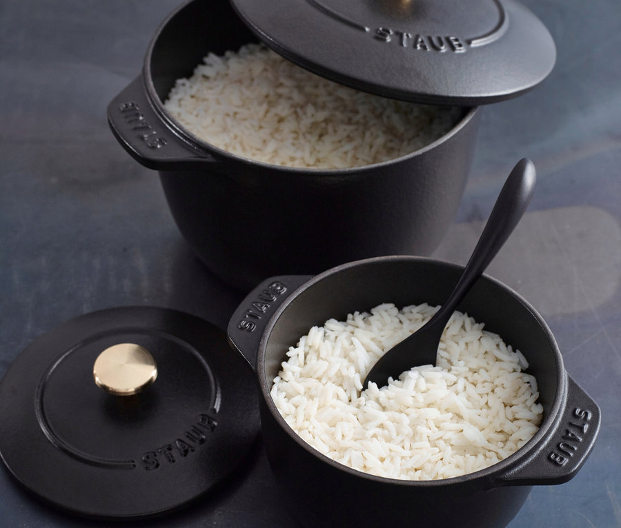 Zwei schwarze Staub Reis-Cocottes, beide gefüllt mit Reis. Eine Cocotte hat einen Löffel darin und einen Deckel daneben.