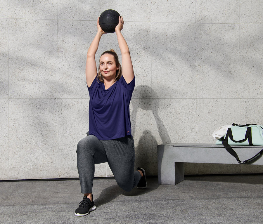 Frau macht Ausfallschritte und hält einen Medizinball über den Kopf, sie trägt eine Trainingshose und ein violettes Oversize-Sportshirt.