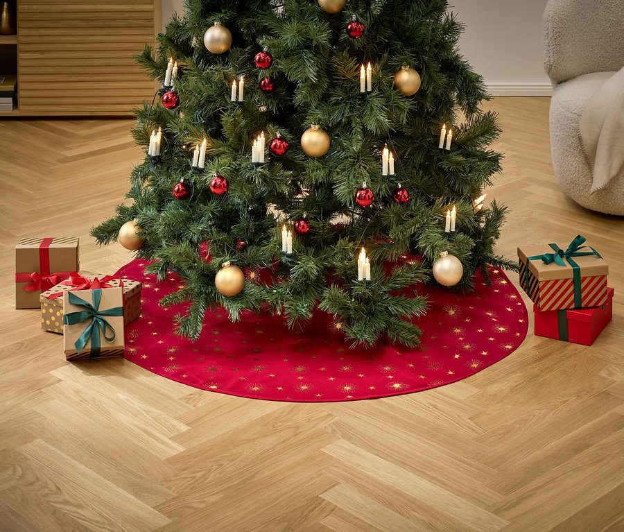 Ein Weihnachtsbaum geschmückt mit roten und goldenen Ornamenten und Lichtern steht auf einem roten Rock mit goldenen Sternen, umgeben von Geschenken.