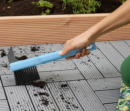 Frau reinigt graue Terrassenplatten mit einer blauen Bürste.