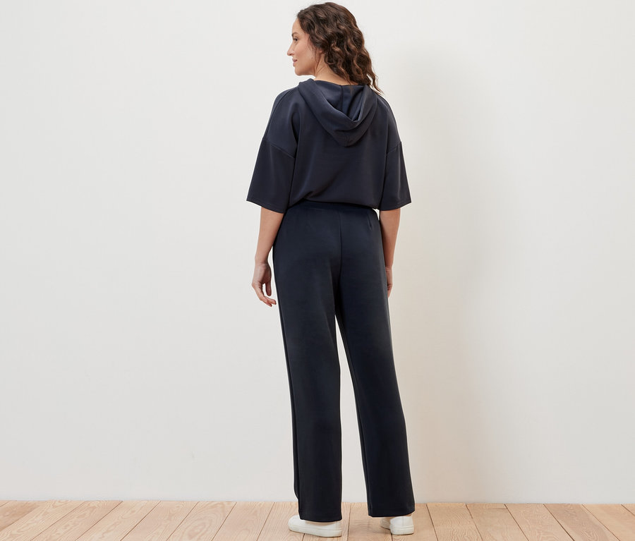 Eine Frau posiert von hinten in Joggpants aus Modal-Mix und einem Kapuzenpullover vor einer cremefarbenen Wand.
