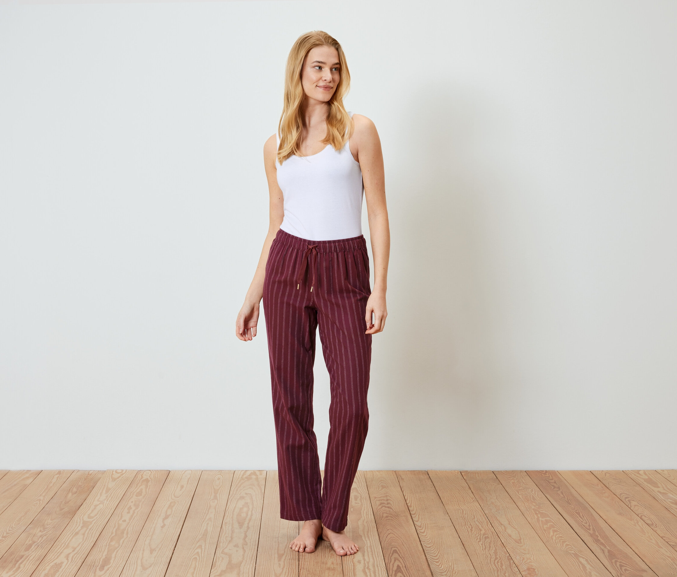 Blonde Frau steht auf einem Holzboden und trägt ein weißes Tanktop und weinrote, gestreifte Hosen.