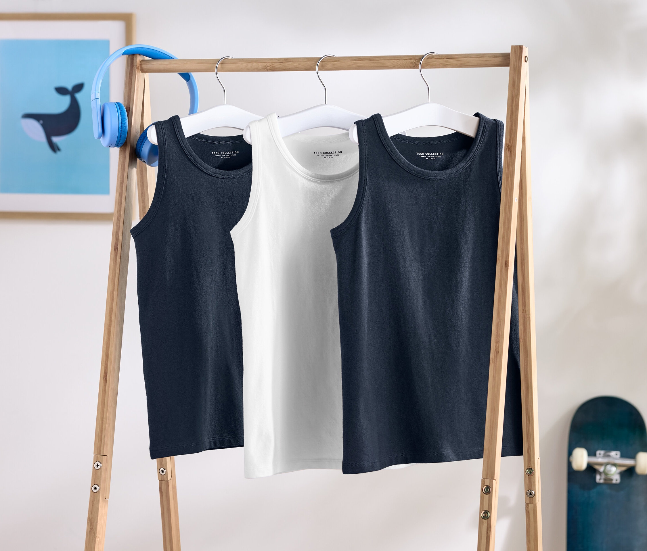 Drei ärmellose Shirts hängen auf einem Holzständer, dahinter ein Skateboard und Kopfhörer.