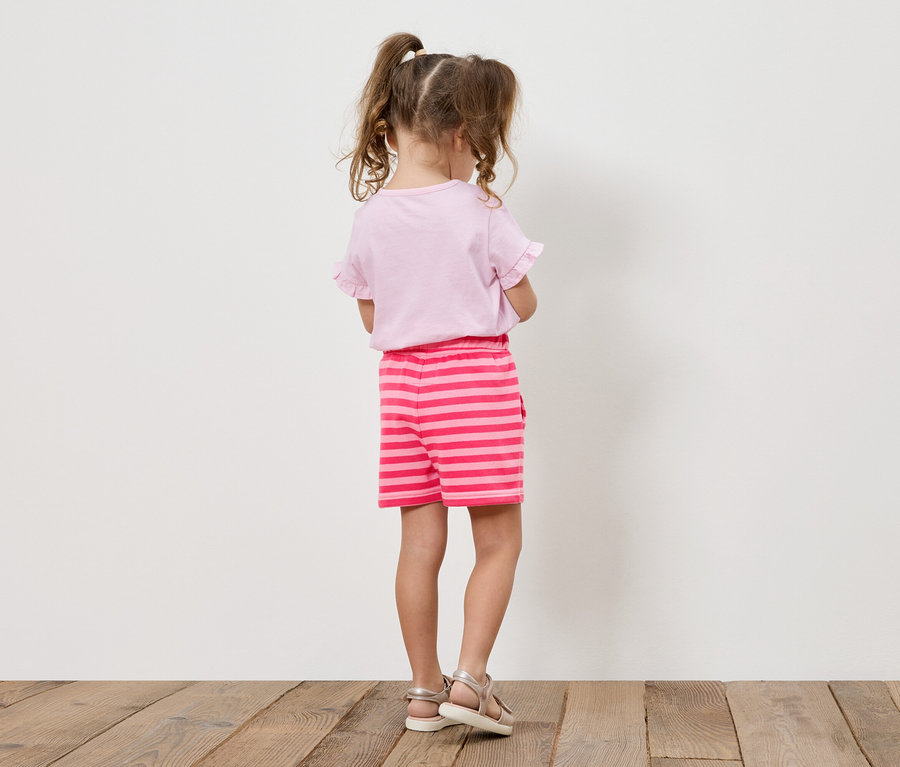 Mädchen mit Zöpfen trägt ein rosa T-Shirt und gestreifte Shorts, von hinten gesehen.