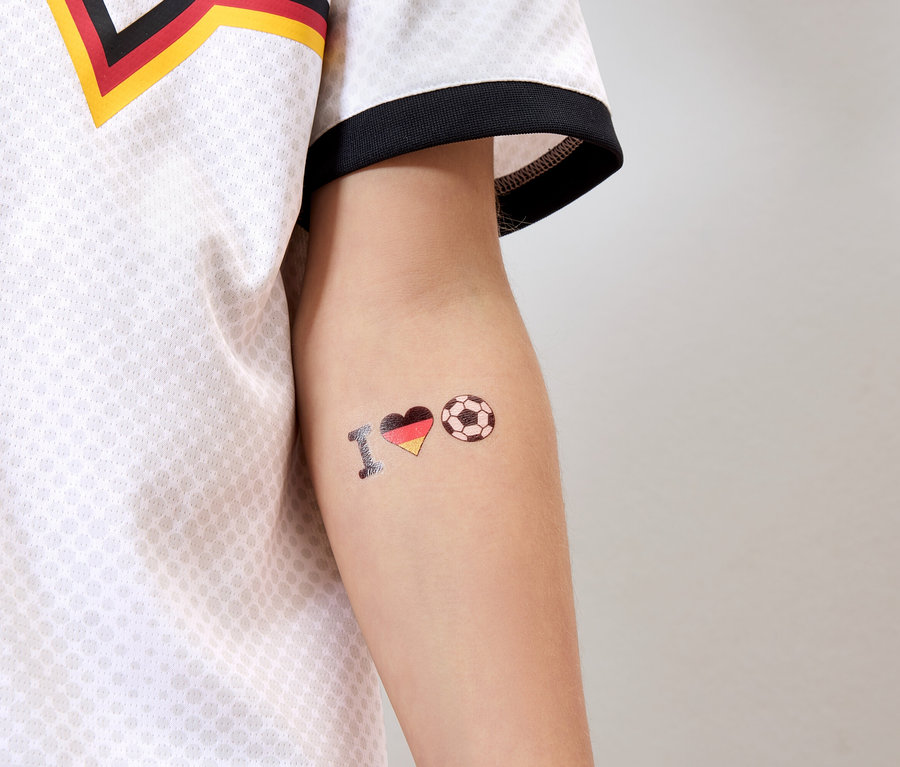 Auf dem Arm einer Person mit Deutschland-Trikot ist ein Tattoo mit der Aufschrift "I <Herz in Deutschlandfarben> Fußball" zu sehen.
