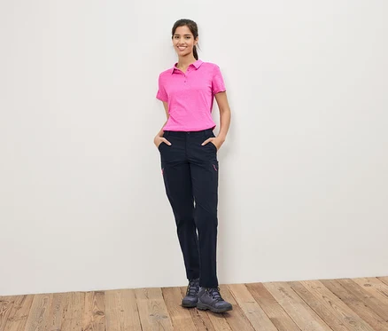 Frau in pinkem Poloshirt und dunkler Hose steht vor einer weißen Wand.