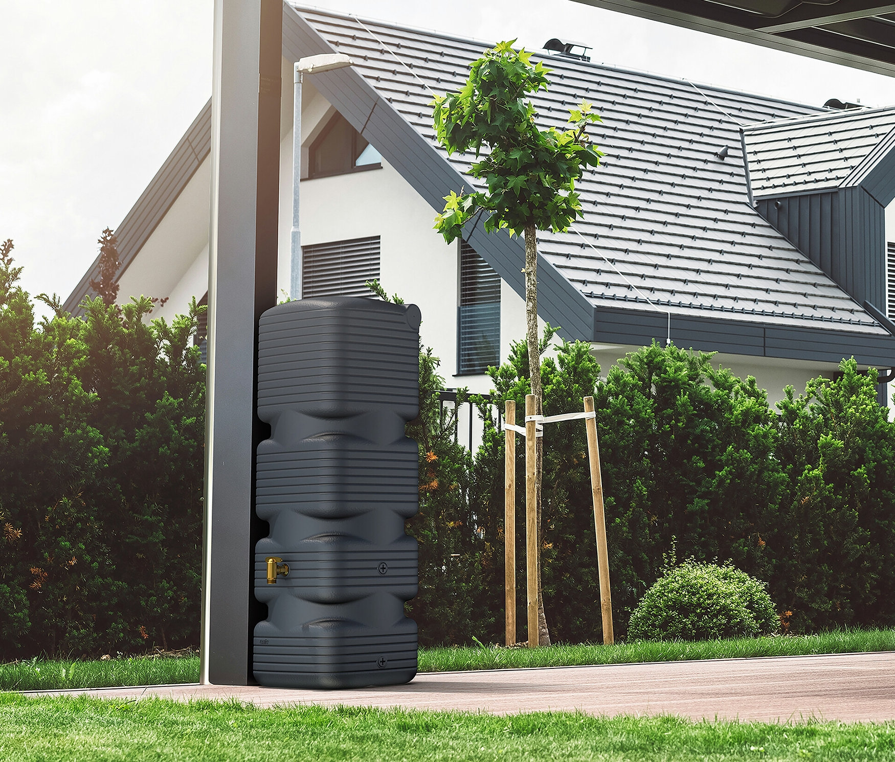 Eine graue Regenwassertonne steht neben einem modernen Haus mit Garten.