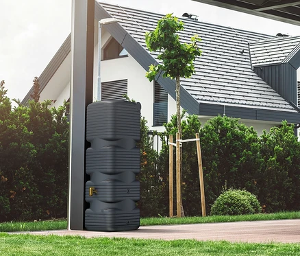 Eine graue Regenwassertonne steht neben einem modernen Haus mit Garten.