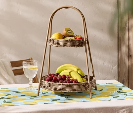 Ein zweistöckiger Obstkorb aus Weide steht auf einem Tisch mit einer Tischdecke mit Zitronenmuster. Auf dem oberen Korb befinden sich eine Zitrone, eine Kiwi und Erdbeeren, während sich auf dem unteren Korb Bananen und rote Trauben befinden.