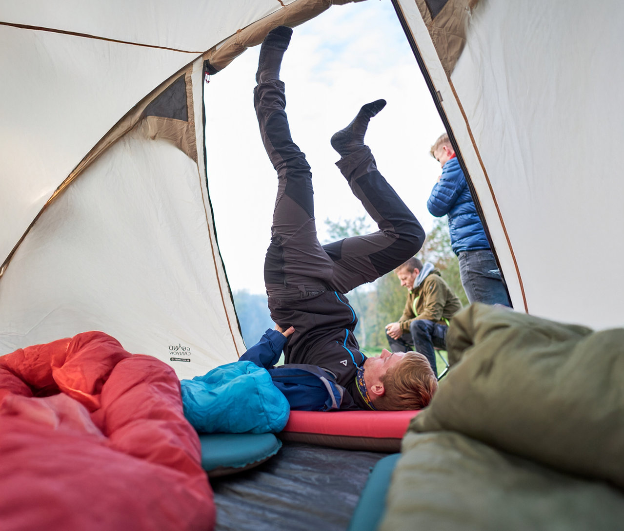 Ein Mann macht einen Kopfstand in einem Zelt, mit Schlafsäcken im Vordergrund und zwei Männern außerhalb des Zeltes.