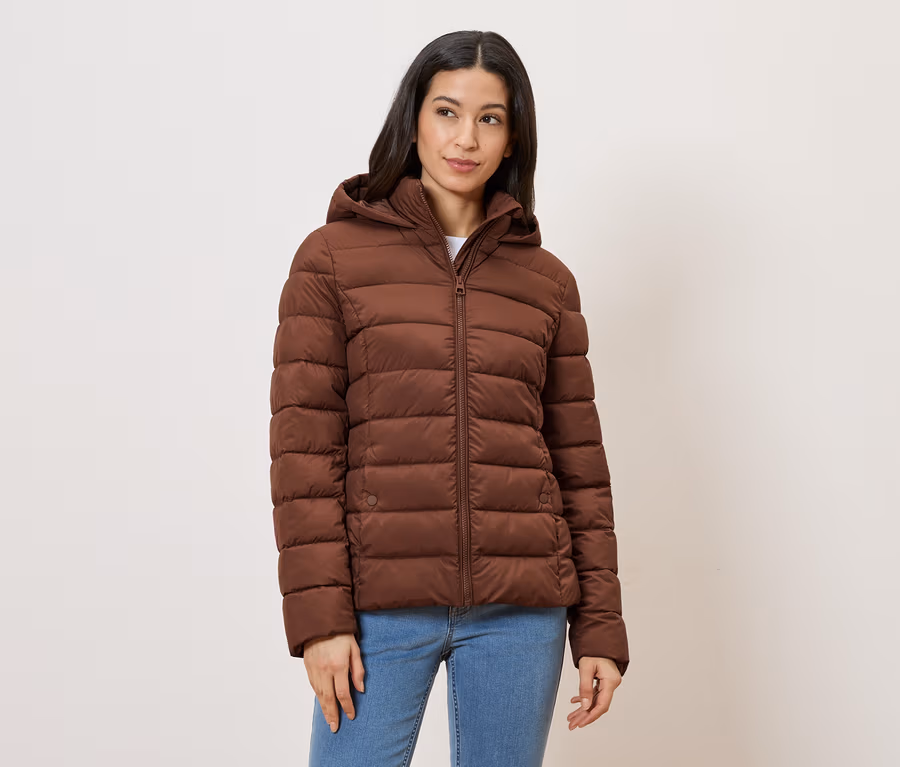 Frau mit Jeans und brauner Steppjacke mit Kapuze vor hellem Hintergrund.