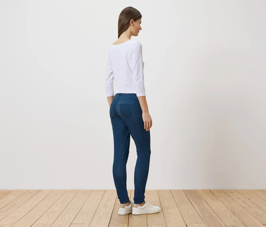 Eine Frau steht seitlich in Jeans, einem weißen Oberteil und weißen Schuhen vor einer weißen Wand und einem Holzboden.