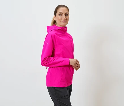 Frau mit Pferdeschwanz trägt pinke Kapuzenjacke und schwarze Hose vor einem weißen Hintergrund.