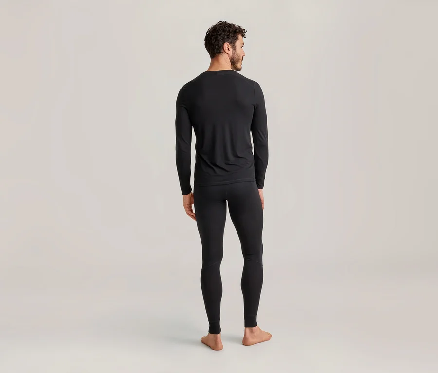 Mann von hinten, der schwarze Thermounterwäsche, bestehend aus einem langärmligen Oberteil und Leggings, vor einem hellen Hintergrund trägt.