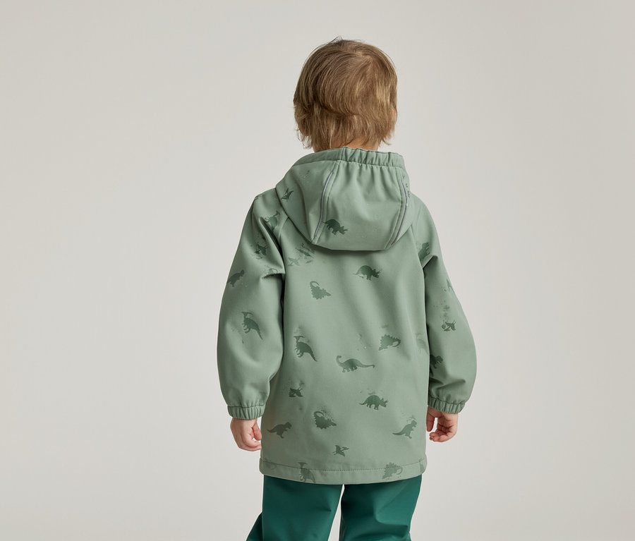 Kind von hinten in grüner Dino-Jacke mit Kapuze und grüner Hose vor hellem Hintergrund.