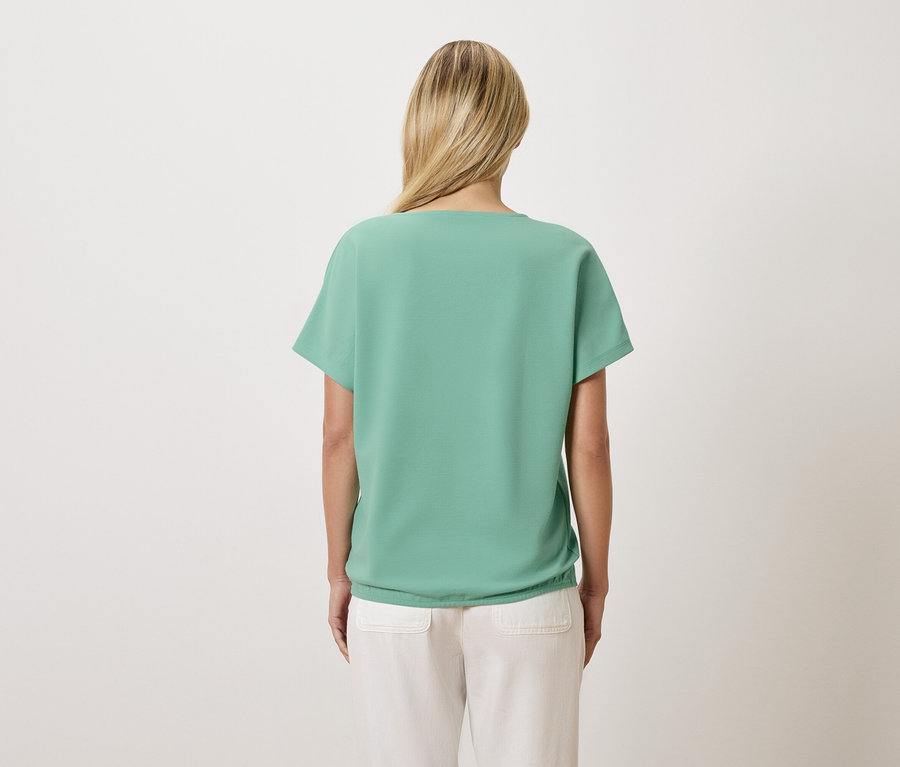 Rückansicht einer Frau mit blonden Haaren, die ein hellgrünes T-Shirt und eine helle Hose trägt.