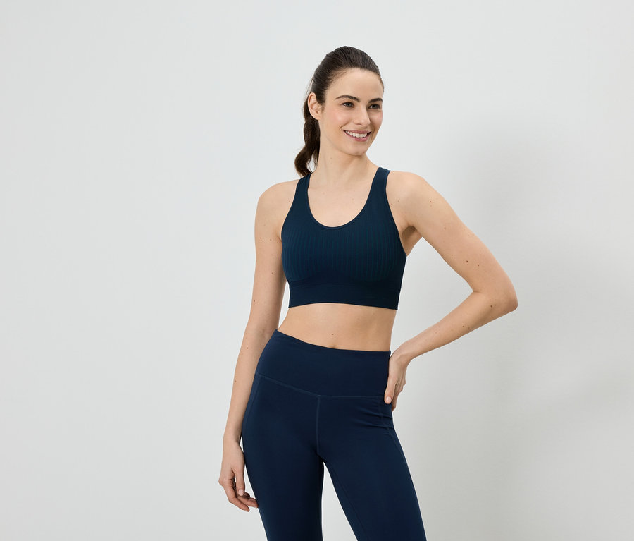 Frau in marineblauem Sport-BH und Leggings, eine Hand an der Hüfte, vor einem weißen Hintergrund.