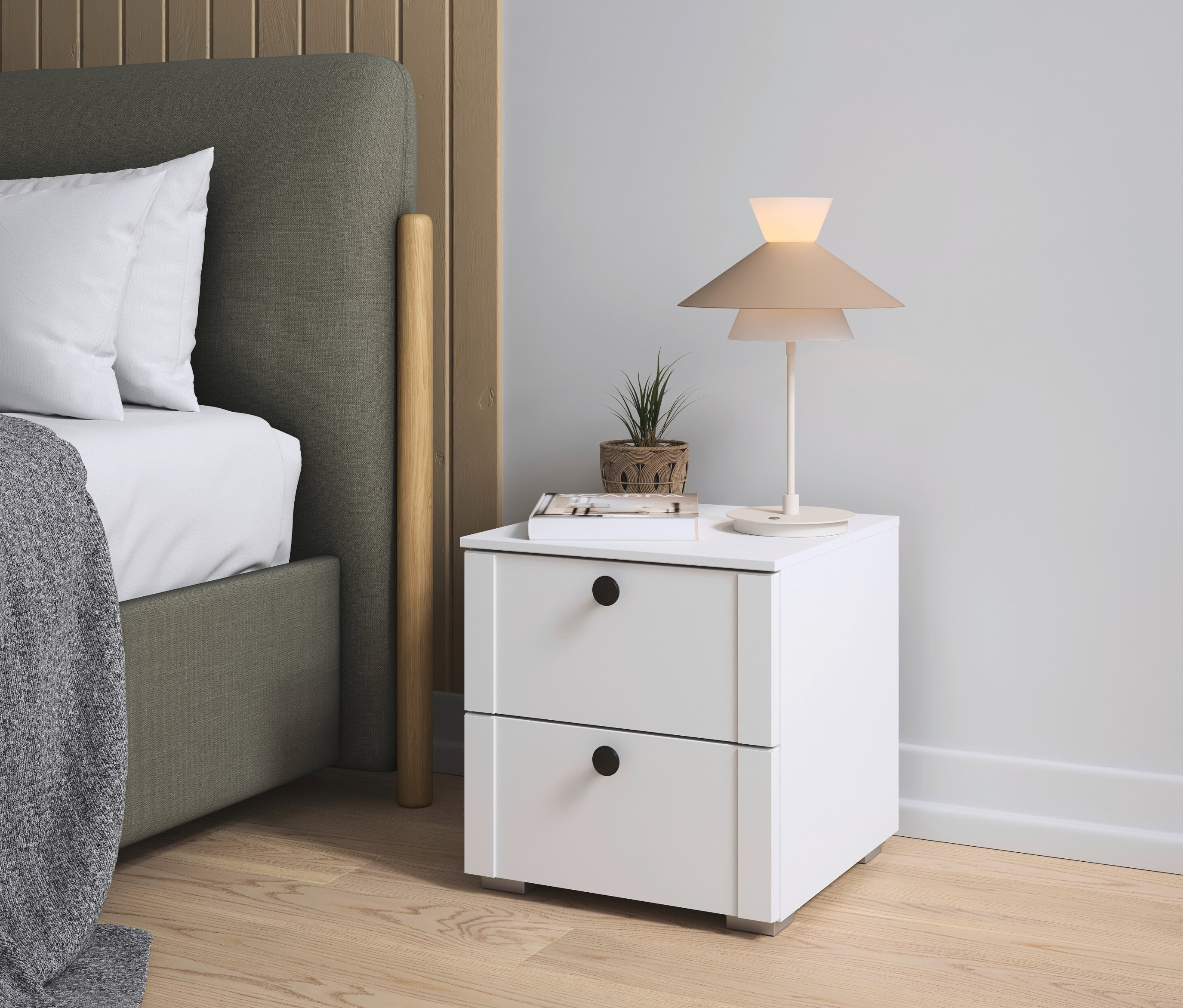 Neben einem Bett steht ein weißer Nachttisch mit zwei Schubladen. Auf dem Tisch stehen eine Lampe, eine Pflanze und ein Buch.