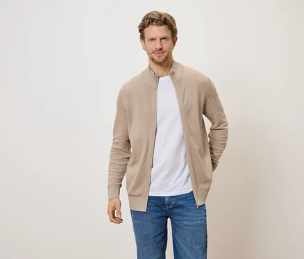 Mann mit hellbraunem Cardigan mit Reißverschluss, weißem T-Shirt und Jeans vor hellem Hintergrund.