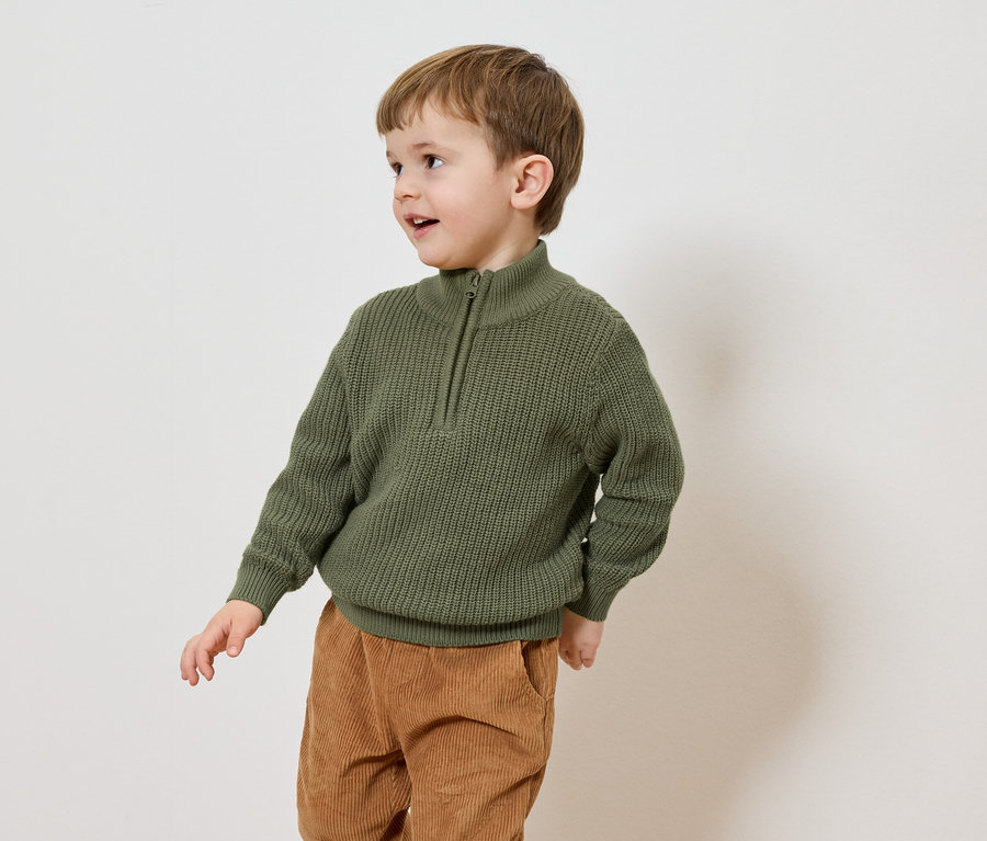Ein Junge mit braunen Haaren trägt einen olivgrünen Strickpullover mit Reißverschluss und braune Cordhosen vor einem weißen Hintergrund.