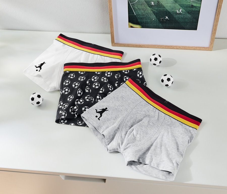 Drei Boxershorts liegen auf einem Tisch, eine weiße, eine graue und eine schwarze mit Fußballmuster.