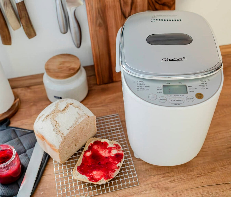 Brotbackautomat neben angeschnittenem Brotlaib und Marmeladenbrot auf einem Holztisch.