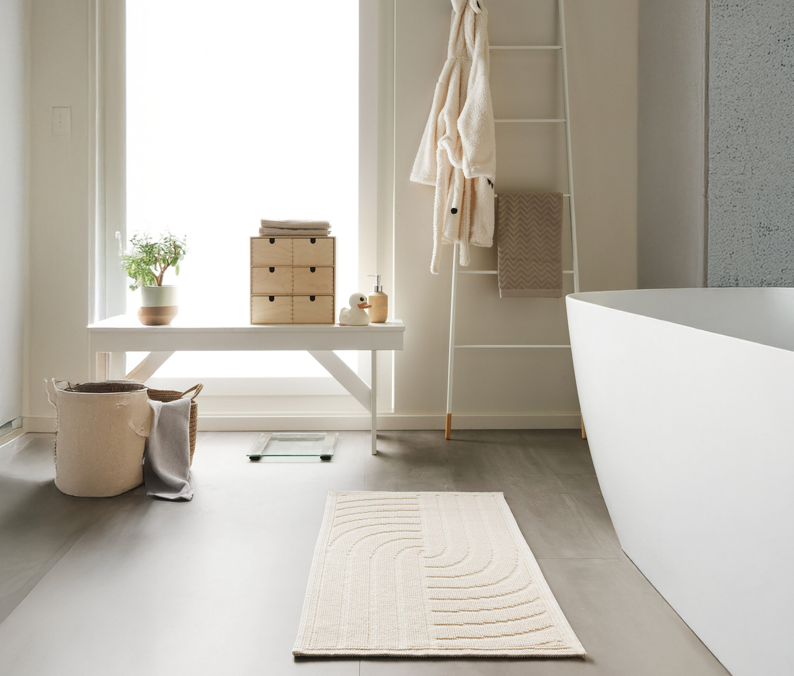 Eine cremefarbene benuta Badematte »Jojo« (50 x 80 cm) mit Wellenmuster liegt auf einem grauen Badezimmerboden.