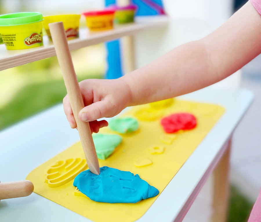 Ein Kind formt blaue Knete mit einem Holzstab auf einer gelben Matte. Im Hintergrund stehen Play-Doh-Dosen auf einem Regal.