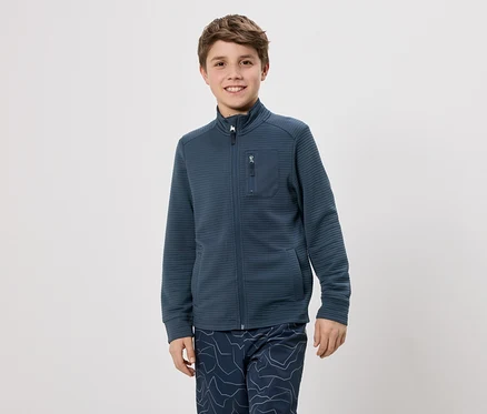 Ein Junge mit braunen Haaren trägt eine dunkelblaue Jacke mit Reißverschluss und eine Hose mit blauem Muster vor einem weißen Hintergrund.