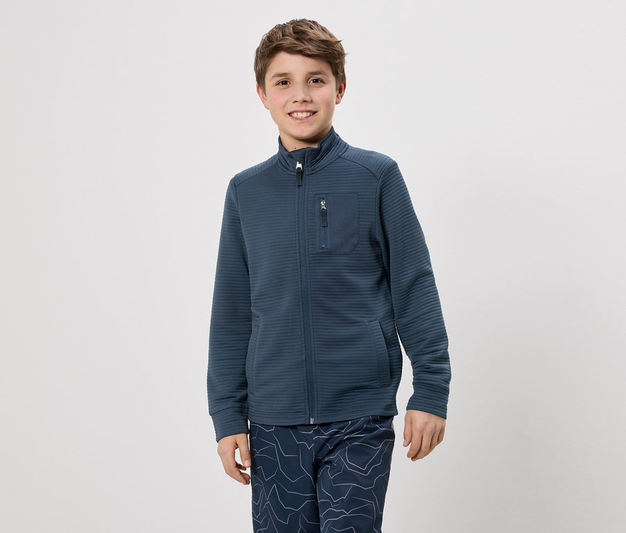 Ein Junge mit braunen Haaren trägt eine dunkelblaue Jacke mit Reißverschluss und eine Hose mit blauem Muster vor einem weißen Hintergrund.