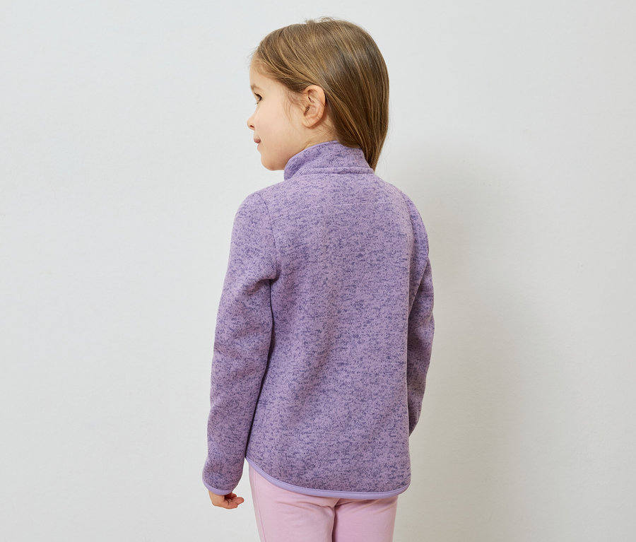 Mädchen von hinten, mit lila Fleecejacke und rosa Hose vor einem weißen Hintergrund.