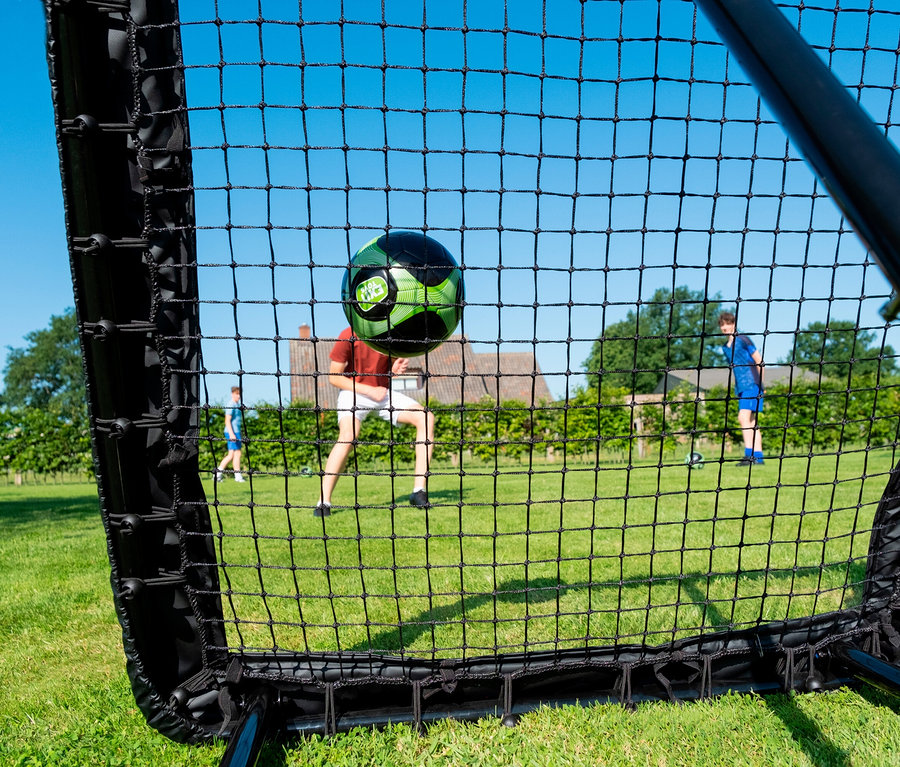 Auf Rasenplatz köpft Mann grünen Ball vor schwarzem Netz, während zwei Jungen im Hintergrund stehen.