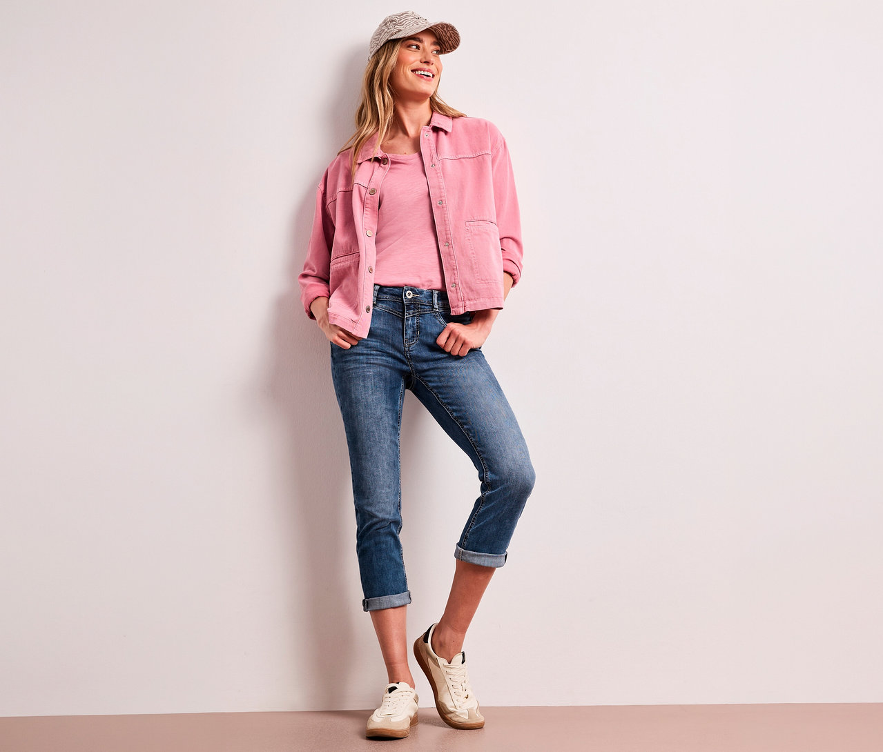 Lächelnde Frau trägt eine gemusterte Kappe, eine rosa Jacke, ein rosa T-Shirt und blaue Street One 7/8-Jeans »Jane« mit umgeschlagenen Säumen, an eine weiße Wand gelehnt.