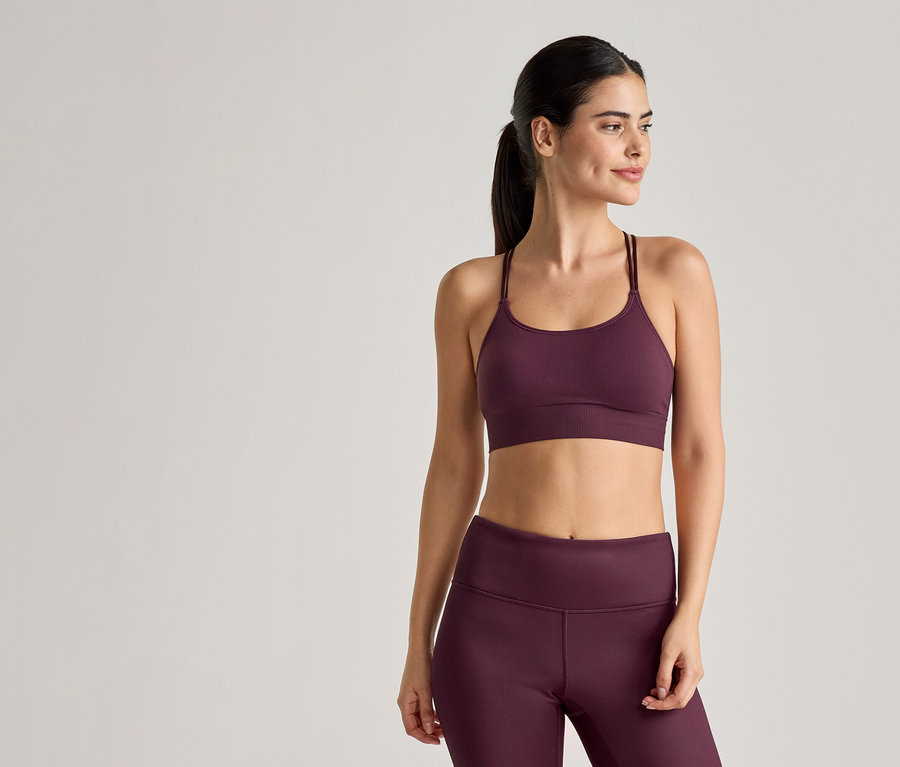 Frau in Sportbekleidung in Pflaumenfarbe, bestehend aus Sport-BH mit dünnen Trägern und Leggings, posiert vor einem hellen Hintergrund.