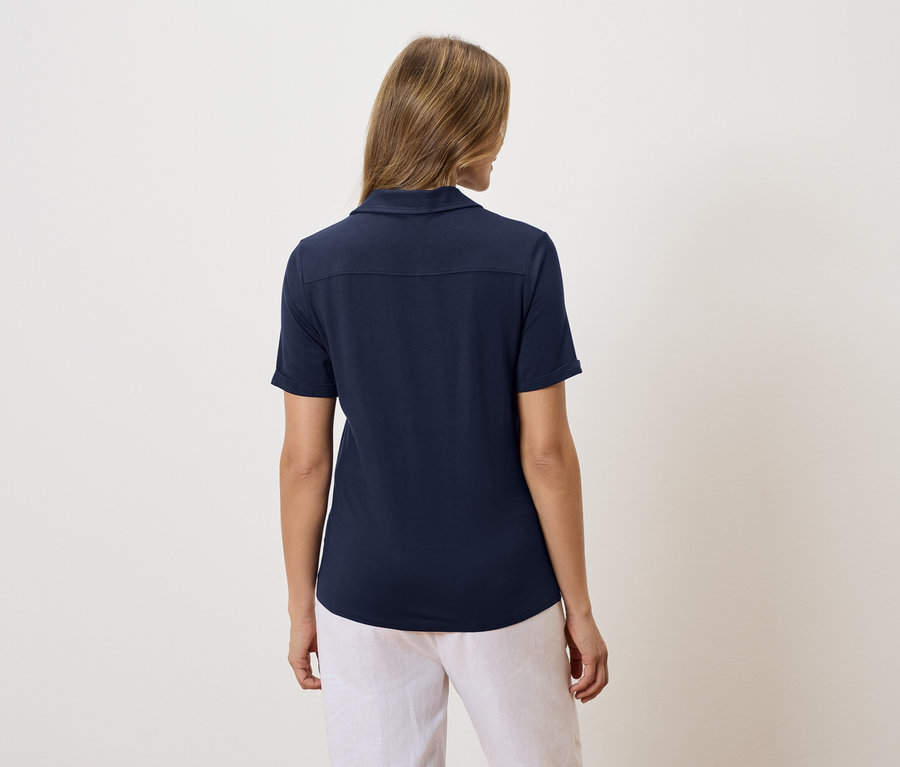 Rückansicht einer Frau mit blonden Haaren, die ein marineblaues Poloshirt und eine weiße Hose trägt.