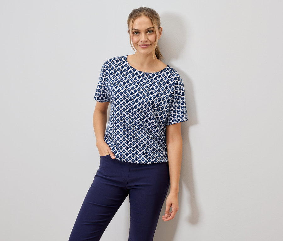 Frau mit blau-weiß gemustertem T-Shirt und blauen Hosen, eine Hand in der Hosentasche.