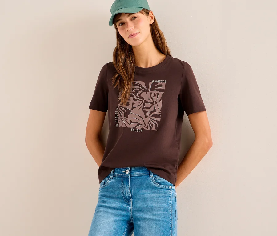 Frau trägt ein braunes CECIL Placement-Shirt mit floralem Muster, hellblaue Jeans und eine grüne Kappe. Sie lehnt an einer hellen Wand.