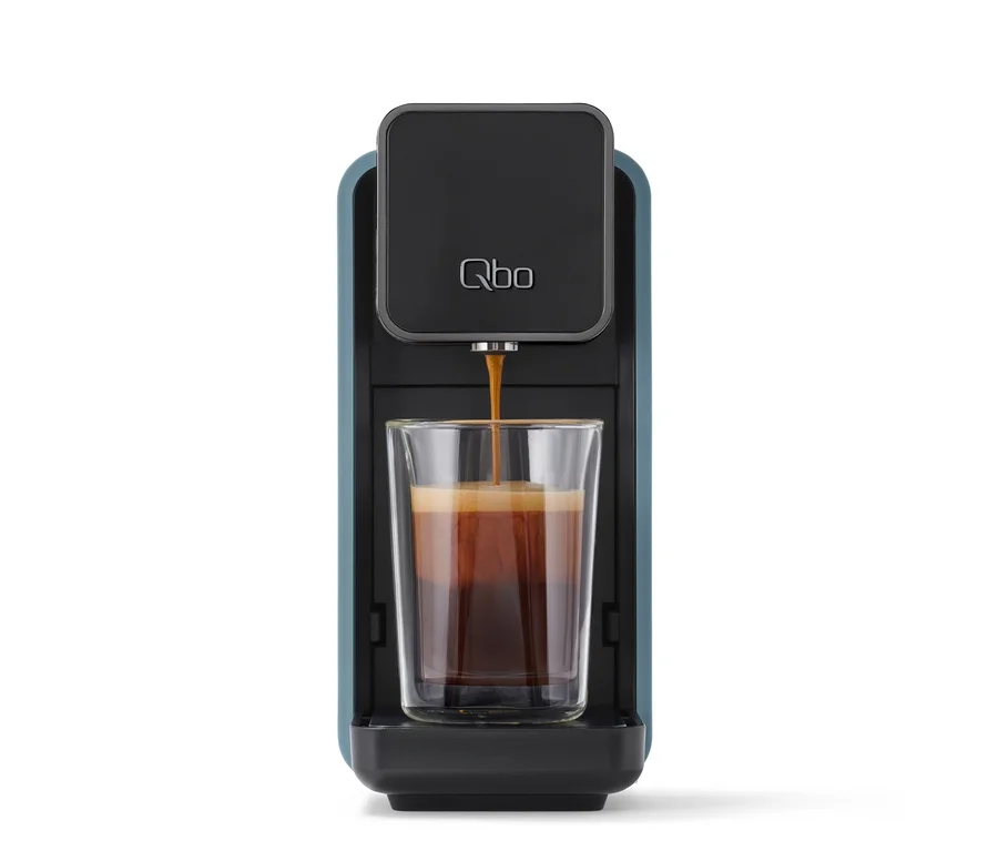 Blaue und schwarze Kaffeemaschine mit Qbo-Logo, die Kaffee in ein doppelwandiges Glas füllt, das halb voll ist.