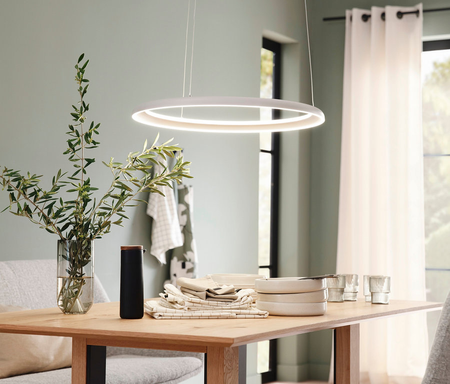 Ein heller Holztisch ist mit Schalen, Gläsern und einem Olivenzweig in einer Vase gedeckt, beleuchtet von einer weißen runden SCHÖNER WOHNEN-Kollektion Pendelleuchte "Quillo" in Sandbrise. Im Hintergrund sind eine hellgrüne Wand und Fenster mit weißen Vorhängen zu sehen.